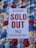 A white sign with bold red text stating 'EVENT SOLD OUT NO PARKING REFUNDS' is placed on a colorful sequined surface composed of blue, red, gold, and silver circular patterns.
