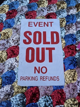 A white sign with bold red text stating 'EVENT SOLD OUT NO PARKING REFUNDS' is placed on a colorful sequined surface composed of blue, red, gold, and silver circular patterns.