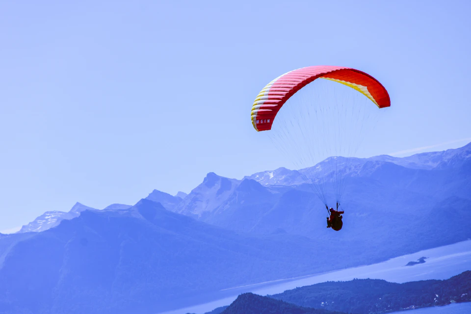 The image depicts a person paragliding over a mountain range, with their body suspended in mid-air and attached to a red and yellow parachute. The paraglider is positioned on the right side of the frame, soaring above the mountains that stretch across the background. The sky forms the backdrop for this thrilling adventure, with its light blue color contrasting beautifully against the vibrant hues of the parachute and mountain range.