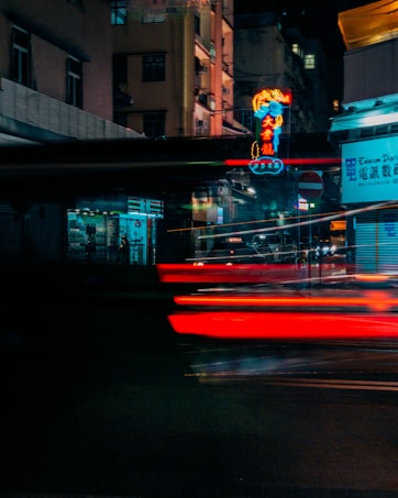 A nighttime urban scene featuring bright, colorful neon signs in a city street, casting vibrant reflections on the surrounding buildings. Blurred red streaks, likely from passing cars, add a dynamic motion effect, while shadows and dim lighting create a sense of depth and intrigue.