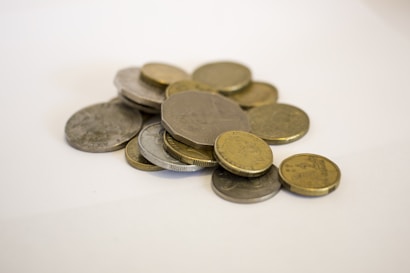 A small pile of mixed coins lies on a light surface. Various shapes and sizes are visible, including round and multi-sided coins. The coins appear to have different metallic finishes, with both silver and gold tones present.