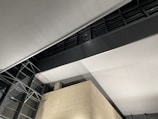 Corrugated ceiling panels with uniform grooves dominate the interior view, intersected by a large dark gray beam. Glass walls with metal frame structures are visible, and a beige tiled wall adds contrast to the modern architecture.