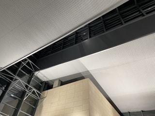 Corrugated ceiling panels with uniform grooves dominate the interior view, intersected by a large dark gray beam. Glass walls with metal frame structures are visible, and a beige tiled wall adds contrast to the modern architecture.