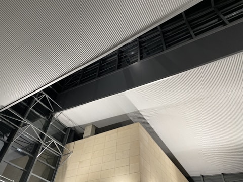 Corrugated ceiling panels with uniform grooves dominate the interior view, intersected by a large dark gray beam. Glass walls with metal frame structures are visible, and a beige tiled wall adds contrast to the modern architecture.