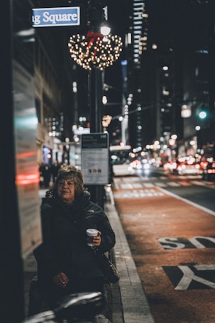A city street scene at night with a woman holding a cup, sitting near a bus stop. The street is busy with traffic and illuminated by streetlights and building lights. Holiday decorations, including a wreath with lights, hang from a lamppost.
