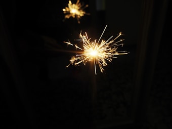 A bright sparkler emits dazzling sparks against a dark background, capturing a sense of celebration and festivity. The light creates a warm glow, casting reflections.