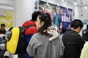 A woman is standing in a crowded indoor space with a cat resting on her shoulder. The cat has a white and gray coat and looks alert. She is wearing a gray shirt and is surrounded by other people. One person is wearing a jacket with a prominent yellow backpack, which has a transparent dome, possibly a pet carrier. The background includes blurred advertisements and lights.