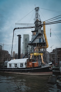 A historical steamship is docked in an urban harbor, featuring a prominent crane and industrial equipment. The ship has a mix of classic and modern elements, with a tent-like structure on its deck and the name 'VOLHARDING 1' visible on its side. In the background, modern high-rise buildings and construction cranes contribute to the city's skyline.