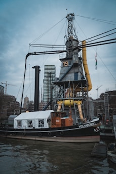 A historical steamship is docked in an urban harbor, featuring a prominent crane and industrial equipment. The ship has a mix of classic and modern elements, with a tent-like structure on its deck and the name 'VOLHARDING 1' visible on its side. In the background, modern high-rise buildings and construction cranes contribute to the city's skyline.