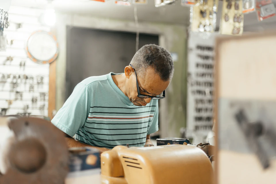 Man works at cluttered workshop desk surrounded by tools and equipment, wearing blue shirt