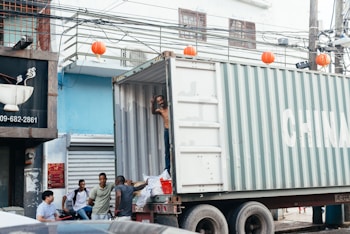 A group of five men interact near a large shipping container labeled with 'CHINA', which is parked on a busy urban street. One man, standing inside the container, is waving or signaling. Red lanterns hang from the wires above the street, while various urban elements such as a partially visible shop sign and closed storefronts are in the background.
