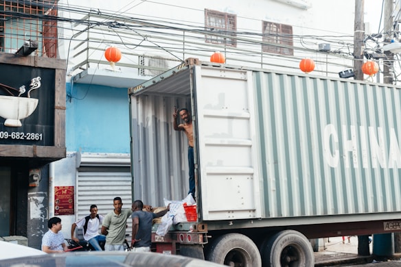 A group of five men interact near a large shipping container labeled with 'CHINA', which is parked on a busy urban street. One man, standing inside the container, is waving or signaling. Red lanterns hang from the wires above the street, while various urban elements such as a partially visible shop sign and closed storefronts are in the background.
