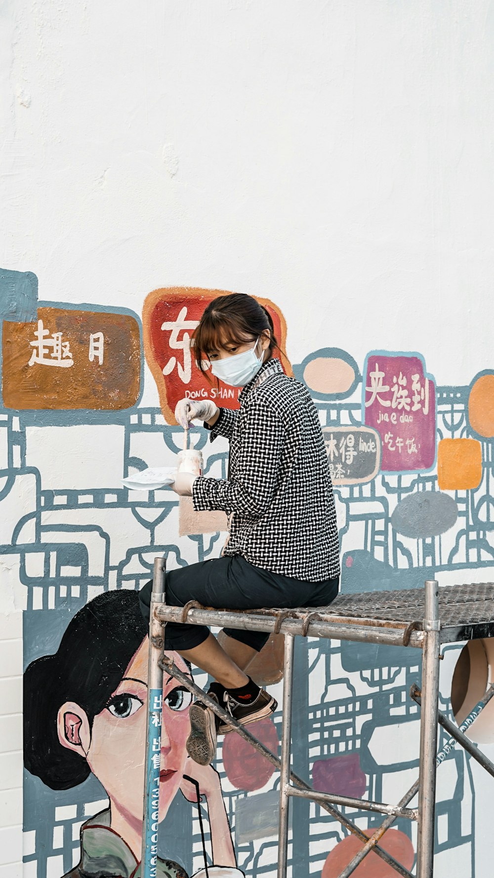 woman wearing white and black tattersall long-sleeved shirt and face mask sitting while holding paint brush