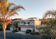 A residential street showcasing a large, elegant house with a beige exterior and white accents, surrounded by lush palm trees. The house features multiple stories with a spacious front yard and a modern design. A black car is parked in the driveway.
