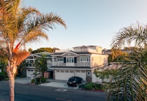 A residential street showcasing a large, elegant house with a beige exterior and white accents, surrounded by lush palm trees. The house features multiple stories with a spacious front yard and a modern design. A black car is parked in the driveway.