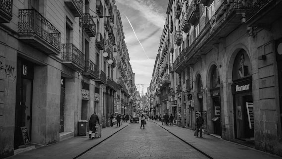 A black and white photo captures a narrow urban street flanked by tall, historic buildings with balconies. The street is paved with cobblestones and features a variety of pedestrians casually strolling along. Some storefronts and signs are visible along the sides, and the sky shows faint contrails from aircraft.