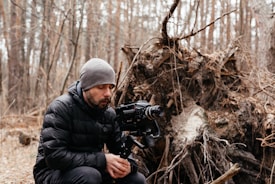 A person in a dark winter jacket and gray beanie is sitting in a forest setting. They are focused on a professional video camera mounted on a tripod. The background features fallen trees and exposed roots, indicating a natural, possibly wild environment.