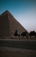 A desert landscape at dusk with the silhouette of a large pyramid in the background. Three camels are walking along a paved road, led by a person wearing a blue top and a headscarf. The scene conveys a sense of ancient history and quietness.