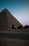 A desert landscape at dusk with the silhouette of a large pyramid in the background. Three camels are walking along a paved road, led by a person wearing a blue top and a headscarf. The scene conveys a sense of ancient history and quietness.