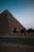 A desert landscape at dusk with the silhouette of a large pyramid in the background. Three camels are walking along a paved road, led by a person wearing a blue top and a headscarf. The scene conveys a sense of ancient history and quietness.