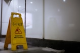 A yellow caution sign is placed on the floor, indicating a wet floor with bilingual text in English and Chinese. The surrounding environment appears to be an indoor space with white walls and a dark floor. A small white cloth is on the ground nearby.