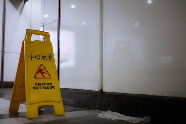 A yellow caution sign is placed on the floor, indicating a wet floor with bilingual text in English and Chinese. The surrounding environment appears to be an indoor space with white walls and a dark floor. A small white cloth is on the ground nearby.