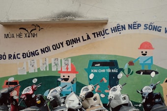 A wall mural features cartoonish characters promoting cleanliness and waste disposal with a bold Vietnamese slogan above it. Several scooters are parked in front of the mural.