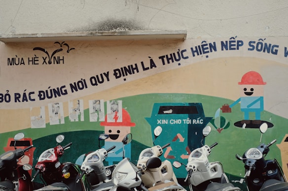 A wall mural features cartoonish characters promoting cleanliness and waste disposal with a bold Vietnamese slogan above it. Several scooters are parked in front of the mural.