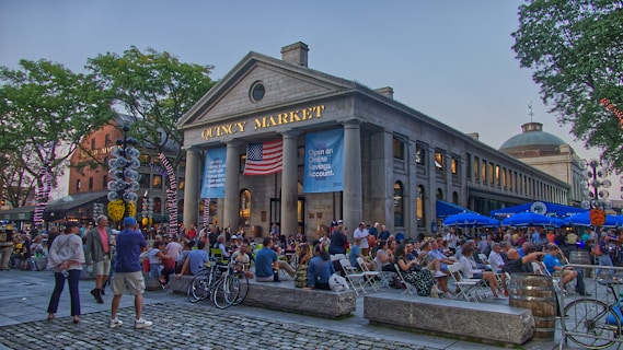 A bustling outdoor scene in front of a historic market building with the words 'Quincy Market' displayed at the top. Numerous people are seated and standing, enjoying the atmosphere, with some trees providing shade. American flags and banners decorate the market, and there are bicycles parked nearby. The scene conveys a lively and communal atmosphere.