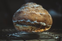 A loaf of rustic bread with a golden-brown crust is sitting on a cooling rack. The surface of the bread has a slightly uneven texture, with flour dusted on top, creating a rustic and homemade appearance. The background is dark, which highlights the bread as the focal point of the image.