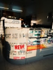 The image depicts an interior of a photo processing or printing shop. Prominent signs and advertisements adorn the counter, including a large 'NEW OFFERS' poster and branding for Kodak Express. Various smaller items and details surround the counter area, including business cards, product brochures, and sample photos. The setting has a warm, sunlight filter casting shadows across the space, highlighting the text and images.