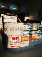 The image depicts an interior of a photo processing or printing shop. Prominent signs and advertisements adorn the counter, including a large 'NEW OFFERS' poster and branding for Kodak Express. Various smaller items and details surround the counter area, including business cards, product brochures, and sample photos. The setting has a warm, sunlight filter casting shadows across the space, highlighting the text and images.