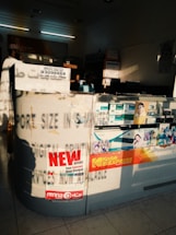 The image depicts an interior of a photo processing or printing shop. Prominent signs and advertisements adorn the counter, including a large 'NEW OFFERS' poster and branding for Kodak Express. Various smaller items and details surround the counter area, including business cards, product brochures, and sample photos. The setting has a warm, sunlight filter casting shadows across the space, highlighting the text and images.