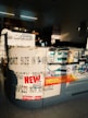 The image depicts an interior of a photo processing or printing shop. Prominent signs and advertisements adorn the counter, including a large 'NEW OFFERS' poster and branding for Kodak Express. Various smaller items and details surround the counter area, including business cards, product brochures, and sample photos. The setting has a warm, sunlight filter casting shadows across the space, highlighting the text and images.