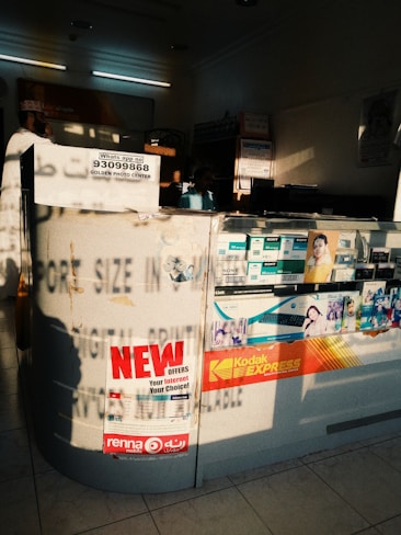 The image depicts an interior of a photo processing or printing shop. Prominent signs and advertisements adorn the counter, including a large 'NEW OFFERS' poster and branding for Kodak Express. Various smaller items and details surround the counter area, including business cards, product brochures, and sample photos. The setting has a warm, sunlight filter casting shadows across the space, highlighting the text and images.