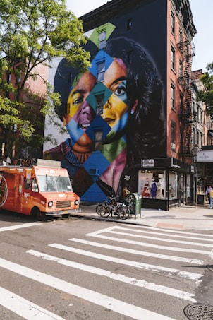 A vibrant mural covers the side of a building, depicting colorful, geometric faces in a patchwork style. In front of the mural, an orange food truck is parked at the corner of an intersection, with bicycles and a small green mailbox nearby. The street has crosswalk markings and is lined with trees.