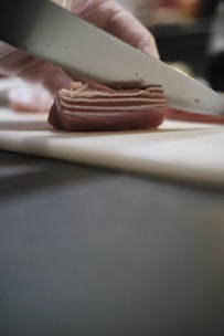 A close-up of a gloved hand slicing a piece of layered cured meat with a sharp knife on a cutting board. The meat displays distinct layers of fat and muscle, indicating it might be bacon or pancetta.