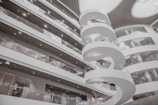 A modern interior featuring a spiral staircase surrounded by multiple levels of open balcony spaces and glass railings. The architectural design is contemporary, with a predominantly white color scheme and rounded structures adding to the aesthetic appeal. Large circular openings in the ceiling allow natural light to illuminate the space.