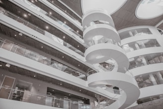 A modern interior featuring a spiral staircase surrounded by multiple levels of open balcony spaces and glass railings. The architectural design is contemporary, with a predominantly white color scheme and rounded structures adding to the aesthetic appeal. Large circular openings in the ceiling allow natural light to illuminate the space.