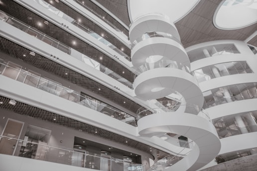 A modern interior featuring a spiral staircase surrounded by multiple levels of open balcony spaces and glass railings. The architectural design is contemporary, with a predominantly white color scheme and rounded structures adding to the aesthetic appeal. Large circular openings in the ceiling allow natural light to illuminate the space.