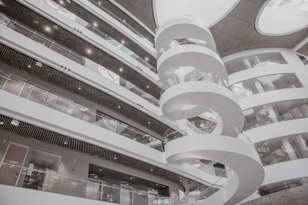 A modern interior featuring a spiral staircase surrounded by multiple levels of open balcony spaces and glass railings. The architectural design is contemporary, with a predominantly white color scheme and rounded structures adding to the aesthetic appeal. Large circular openings in the ceiling allow natural light to illuminate the space.