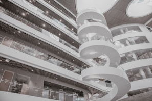 A modern interior featuring a spiral staircase surrounded by multiple levels of open balcony spaces and glass railings. The architectural design is contemporary, with a predominantly white color scheme and rounded structures adding to the aesthetic appeal. Large circular openings in the ceiling allow natural light to illuminate the space.