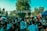 A lively outdoor event with many people gathered under colorful string decorations. The atmosphere is festive, with attendees socializing, and various tents and stalls visible. The setting includes lush greenery with trees and a bright blue sky overhead.