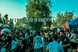 A lively outdoor event with many people gathered under colorful string decorations. The atmosphere is festive, with attendees socializing, and various tents and stalls visible. The setting includes lush greenery with trees and a bright blue sky overhead.