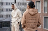 Two individuals are dressed in casual hoodies standing in an urban setting. The person in the foreground wears a tan hoodie with motivational phrases printed on the back. The person in the background leans against a railing wearing a light-colored hoodie with hands in pockets, and there are buildings in the blurry background.
