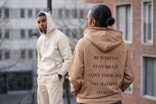 Two individuals are dressed in casual hoodies standing in an urban setting. The person in the foreground wears a tan hoodie with motivational phrases printed on the back. The person in the background leans against a railing wearing a light-colored hoodie with hands in pockets, and there are buildings in the blurry background.