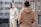 Two individuals are dressed in casual hoodies standing in an urban setting. The person in the foreground wears a tan hoodie with motivational phrases printed on the back. The person in the background leans against a railing wearing a light-colored hoodie with hands in pockets, and there are buildings in the blurry background.