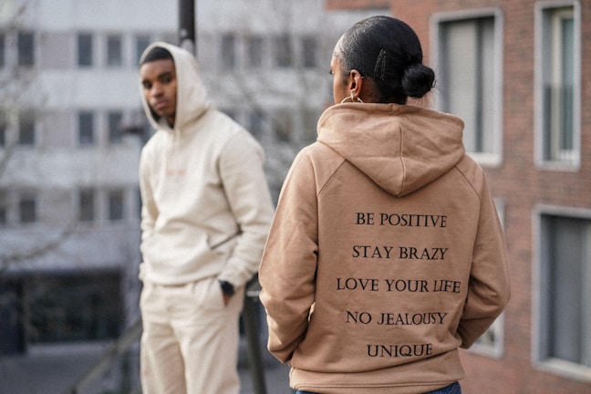 Two individuals are dressed in casual hoodies standing in an urban setting. The person in the foreground wears a tan hoodie with motivational phrases printed on the back. The person in the background leans against a railing wearing a light-colored hoodie with hands in pockets, and there are buildings in the blurry background.
