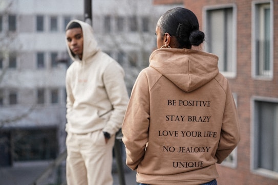 Two individuals are dressed in casual hoodies standing in an urban setting. The person in the foreground wears a tan hoodie with motivational phrases printed on the back. The person in the background leans against a railing wearing a light-colored hoodie with hands in pockets, and there are buildings in the blurry background.