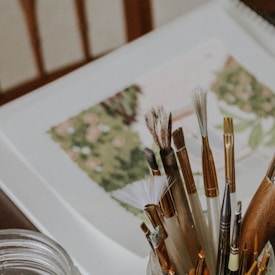 A variety of paintbrushes are arranged in a holder next to an open notebook featuring a floral or nature-themed painting. The scene is focused on the tools of an artist with a soft, warm tone.